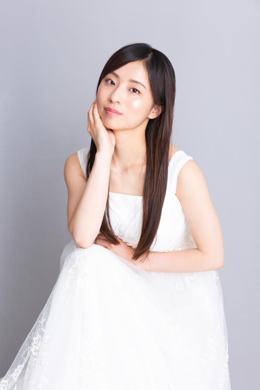 髪の長い白い服を着た女性が頬杖をついている。小嶋早恵写真。