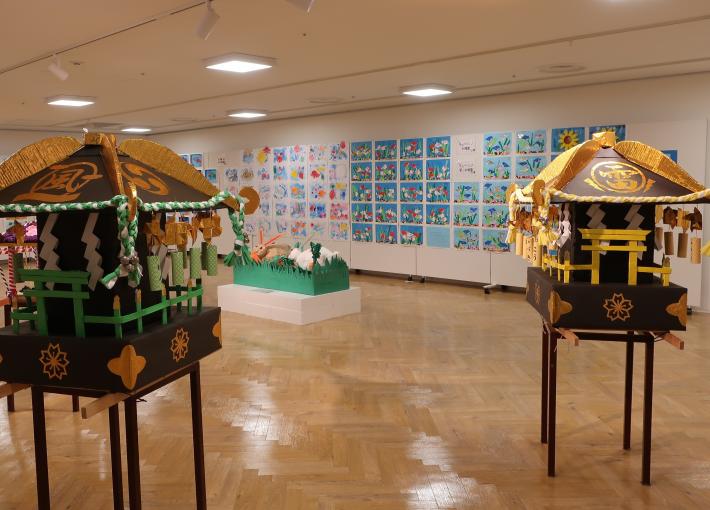 壁に児童絵が貼付されて、床にミニ御おみこし2台とその奥に立体作品が1つ展示されている様子の画像。