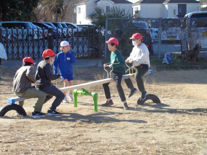 校庭にあるシーソーで遊ぶ子どもたち。2人ずつ左・右にのっており、真ん中で見ている子どもが1人いる。