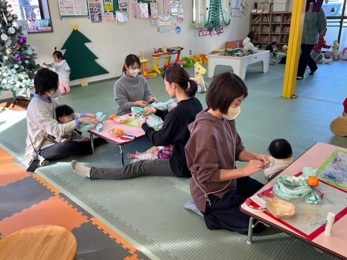 母たちが正月飾りを作成しているところを子が膝の上から見ている写真