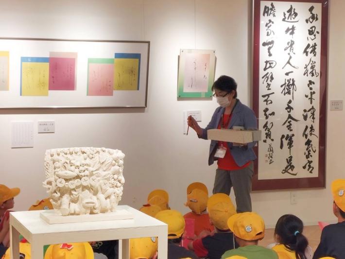 壁に貼られた絵画を見ながら芸術家の解説を聞く小学生たち