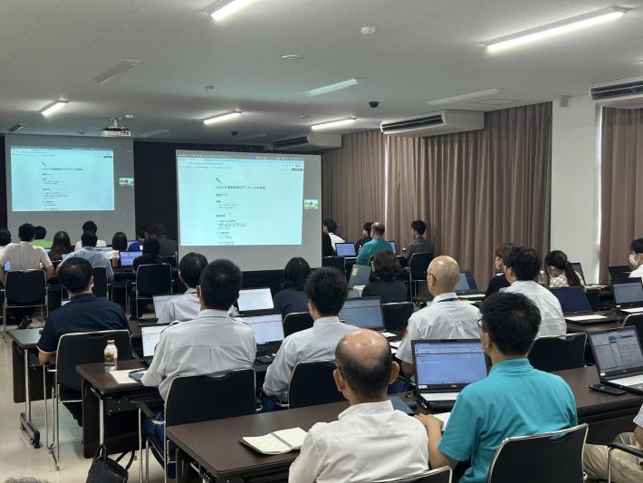 会場の前方に設置されたスクリーンを座って見る職員たち
