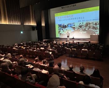 大型のスクリーンを背景に壇上の下に座る参加者にあいさつをする男性