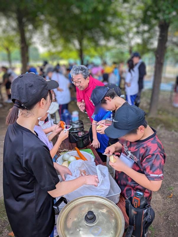 ピンク色のポロシャツを着た男性が子どもたちが野菜の皮をむいている様子を見ている