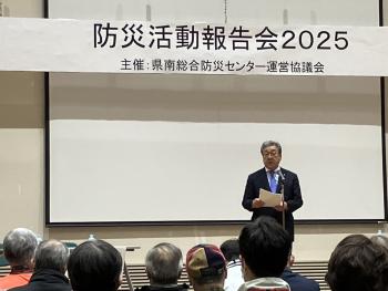 防災活動報告会2025と書かれた看板の下から挨拶をする男性