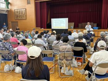 体育館内に集まった着座の参加者に向けてあいさつをする男性