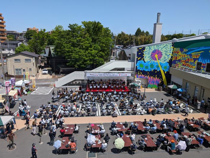 ステージ上とステージ下にいる演者が楽器を演奏している。観客は、椅子に座って演奏を楽しんでいる。