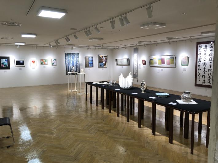 会場の壁に絵画などが展示されている。中央には、工芸などの展示物がある。