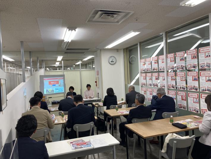 会議室にスーツを着た男性と女性合計10人がこちらに背を向けて座り、その奥にスーツを着た女性2人が座っている