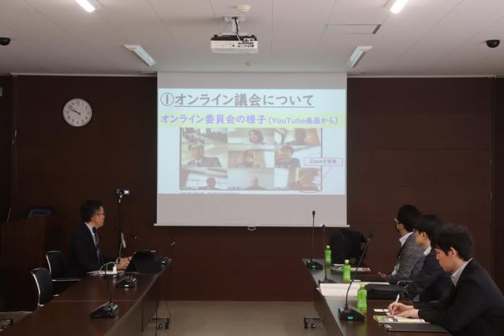 会議室で男性3人が椅子に座ってスクリーンに投影された映像を見ている