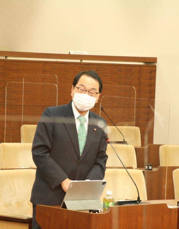 議場にて紺色のスーツを着た男性議員が起立してあいさつをしている。