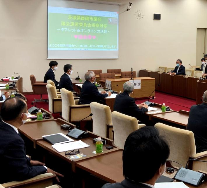 議場のスクリーンに資料が映っている。鹿嶋市議が議席に着席し、スクリーンの資料を見ている写真。