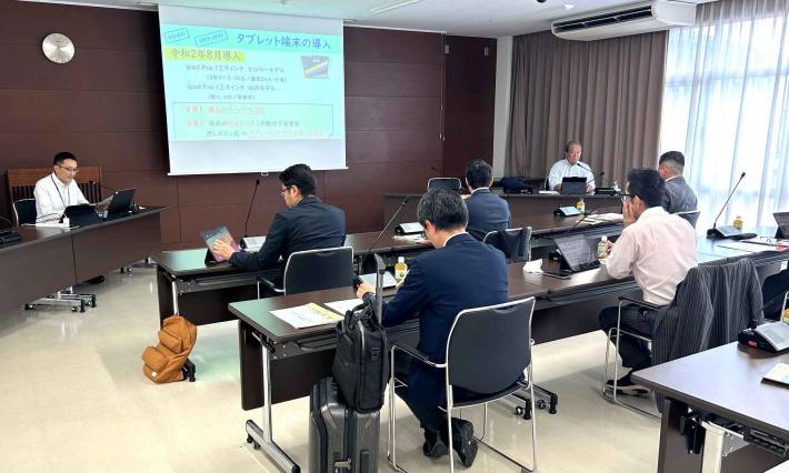 福岡県糸島市議会の議員らが前方のスクリーンに投影された資料を見ながら説明を受けている様子