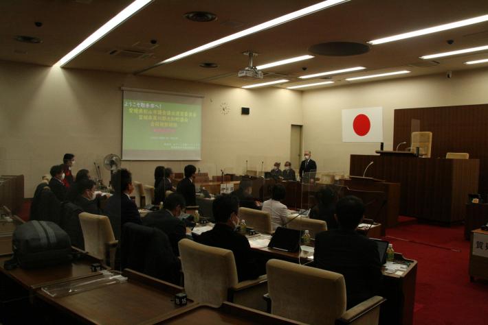 議場後方から移した写真。スクリーンに緑色の資料が映る。参加者は前を向いて座っている。