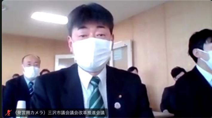 Zoom画面に会議室が映っており、手前に黒いスーツを着た短髪の男性が座っている