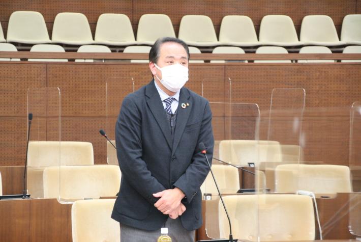 議場にて紺色のスーツを着た男性が起立してあいさつをしている。