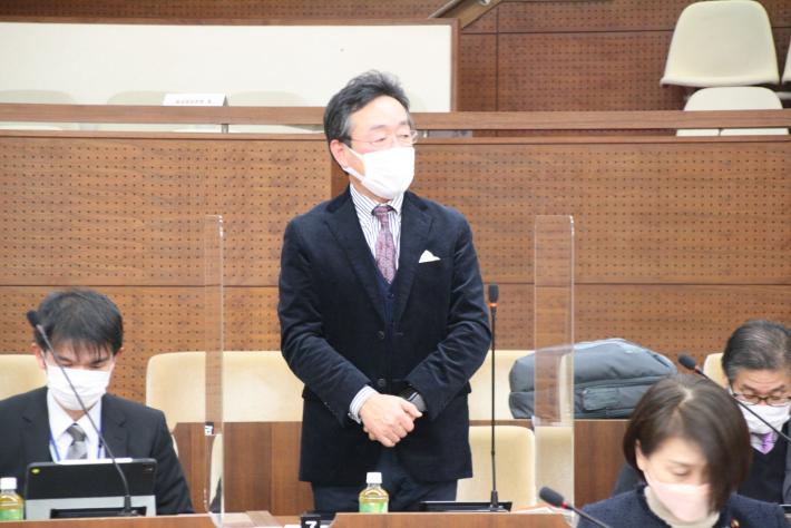 議場の議席にて紺色のスーツを着た男性があいさつをしている。