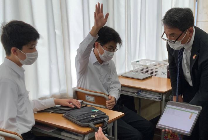 中学校の教室で、タブレット端末を持ちながら議員と生徒が対話している