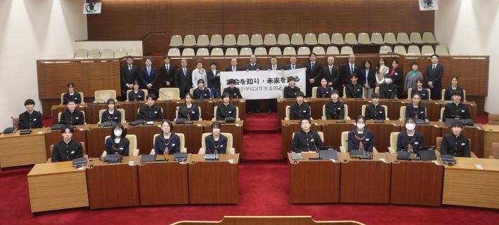 議場にたくさんの生徒が座り、その奥に議員が一列に並んで、笑顔でこちらを向いている