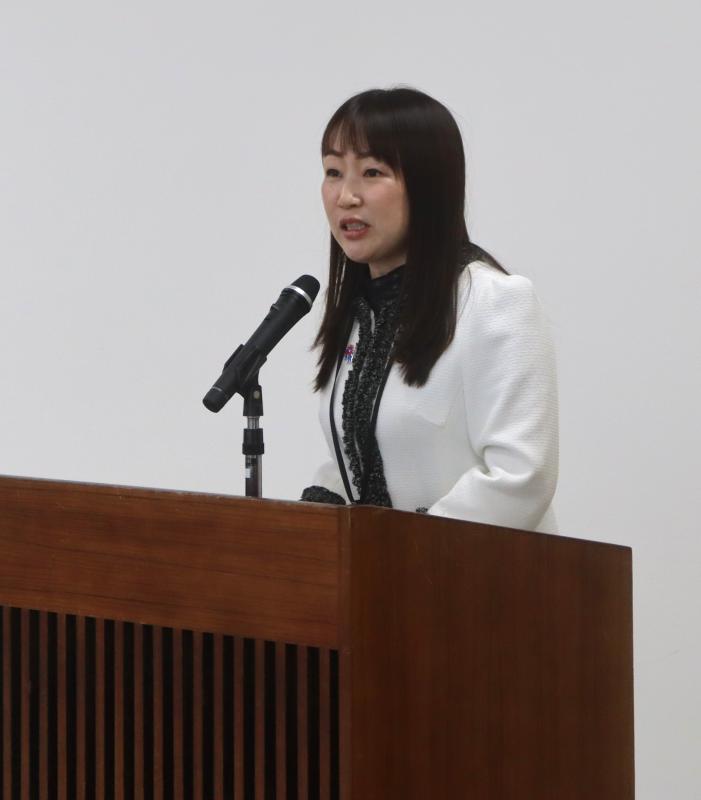 大会議室の演台で白色の上着を着た女性があいさつをしている