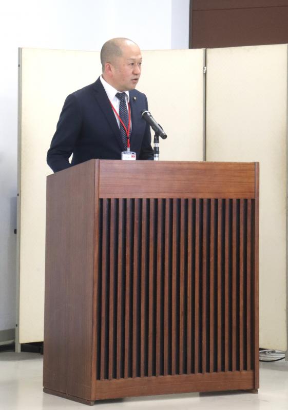 大会議室の演台で黒色の上着を着た男性があいさつをしている