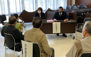 大きな窓のある会議室で、オールバックの男性とスーツを着た短髪の女性、身を乗り出す女性1人と男性3人が向かい合って話している