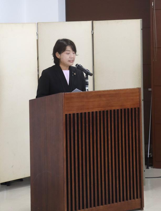 大会議室の演台で黒色の上着を着た女性がが司会をしている
