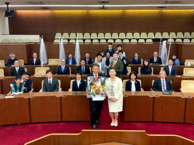 議場で受賞の記念撮影をしている。議員は議席に議長と事務局長が最前中央で一緒に花束を持っている