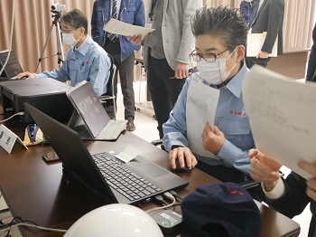 机が2つ置かれ、水色の作業服を着た短髪の女性と男性が着席し、机上のパソコンを操作している