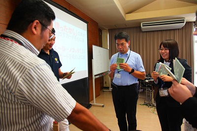 司会の議会事務局職員が、男性議員と女性職員の出したカードを持っている。男性議員がプレゼンを行っている