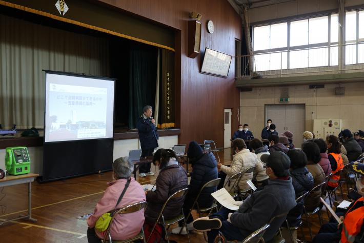 体育館ステージ前にて挨拶をする市長。市長の前にはパイプ椅子に座る多くの人々市長の後ろにはスクリーン