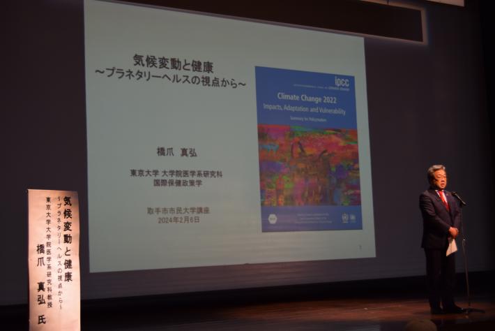 ステージ上で挨拶をする市長。背景にはプロジェクターで映し出されるタイトル「気候変動と健康」