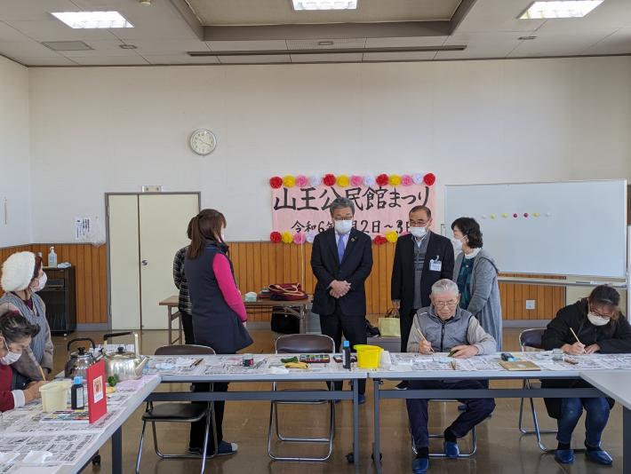 会議室にて椅子と机が並べられ作業をする人々。前方には参加者と話をする市長。背景には「山王公民館まつり」の横看板