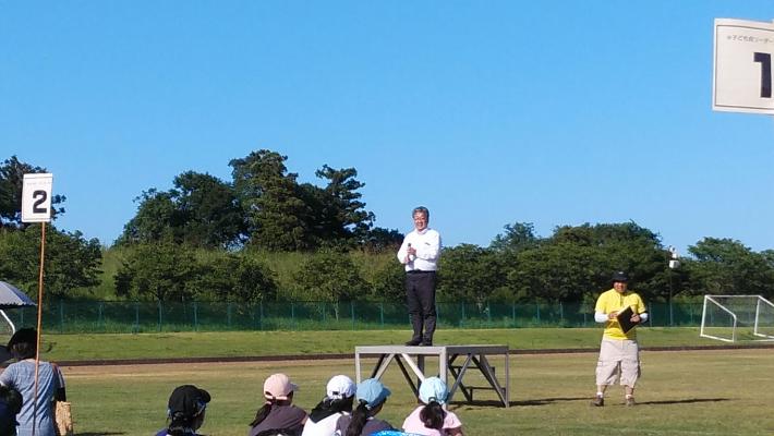 子ども会サマーキャンプのようす。グラウンド様の場所で、青空のもと、マイクを手に挨拶する男性とそれを対面で聞いている子どもがいる