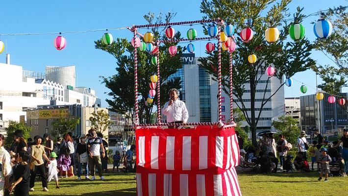 ウェルネスプラザ納涼祭のようす。周囲を提灯で飾った紅白幕つきやぐらの上で挨拶している男性の画像。