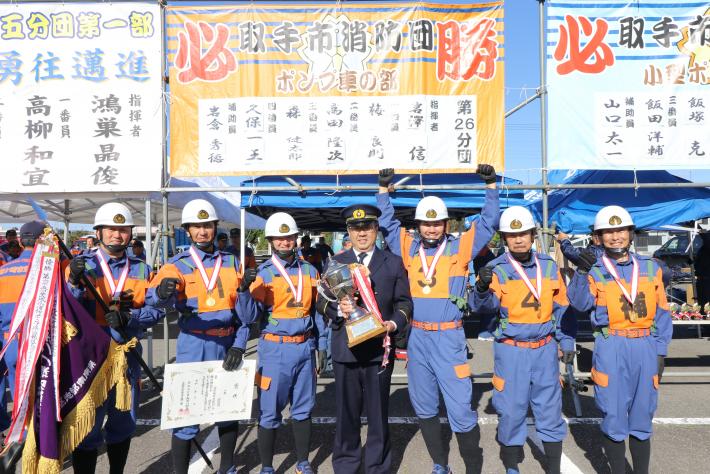 横断幕の前で消防服を着た6人の団員とトロフィーを持った市長が立っている