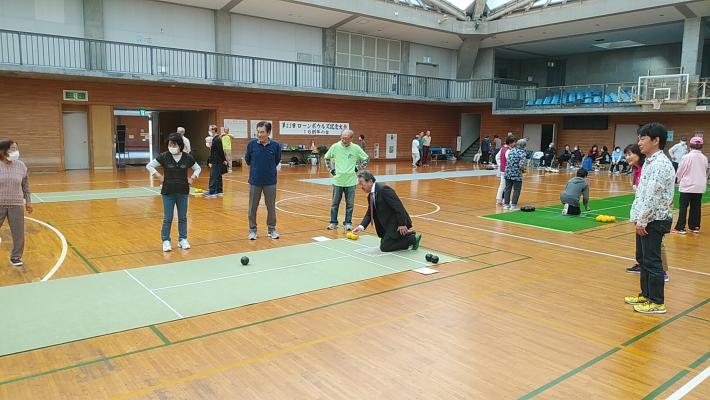 体育館にてローンボウルズをする人々。シートが敷かれたコースの上をボールを転がす市長と見守る市民