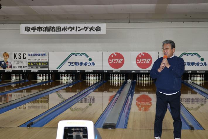 ボウリング場にてレーンを背にあいさつをする市長。レーン上には「取手市消防団…」の横看板」