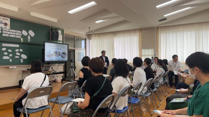 山王小学校学校説明会の様子。教室内に並んだパイプ椅子に座ったひとたちが前方で話す男性の話を聞いている。