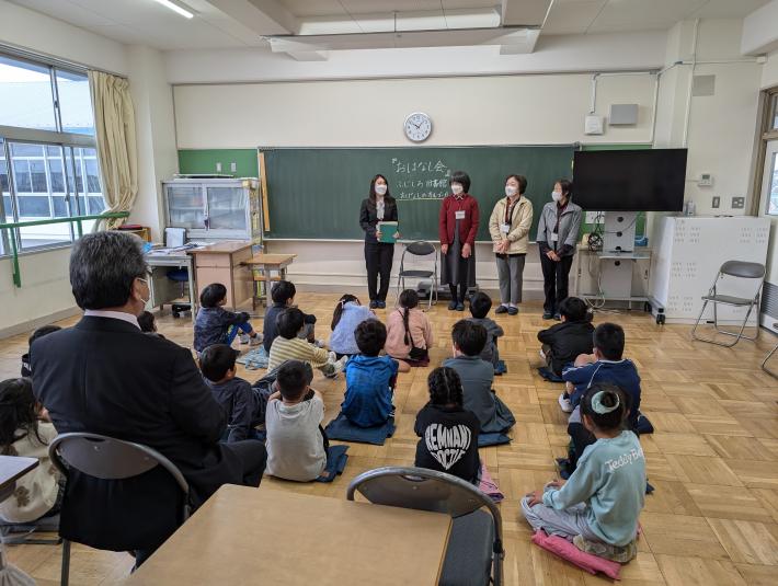 座って黒板の前に立つ女性の話を聞く子どもたちと男性