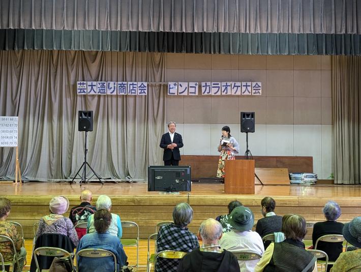 芸大通り商店会ビンゴカラオケ大会と書かれた看板の下からあいさつをする男性