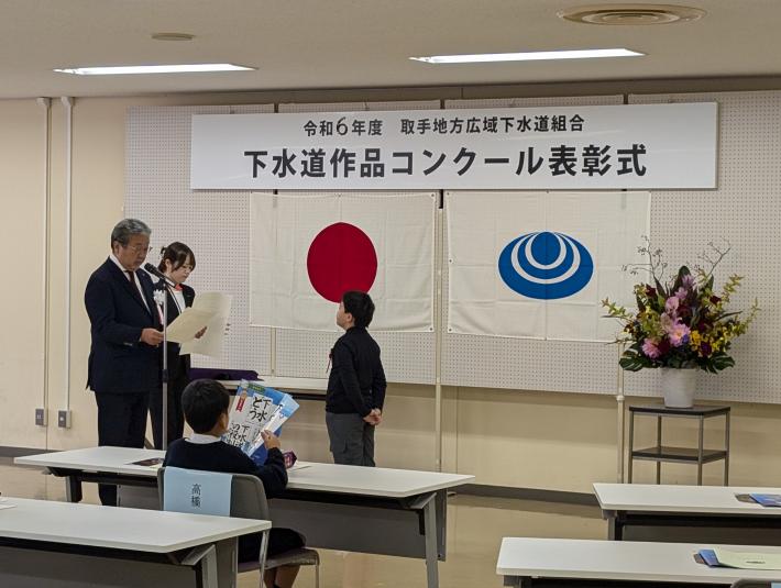 令和6年度取手地方広域下水道組合下水道作品コンクール表彰式と書かれた看板の下で、表彰状を授与する男性