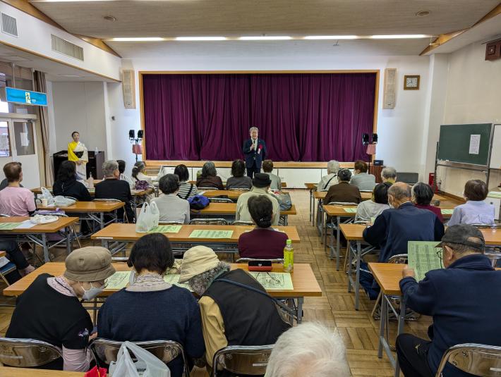 藤色の幕がかかったステージの前からあいさつをする男性