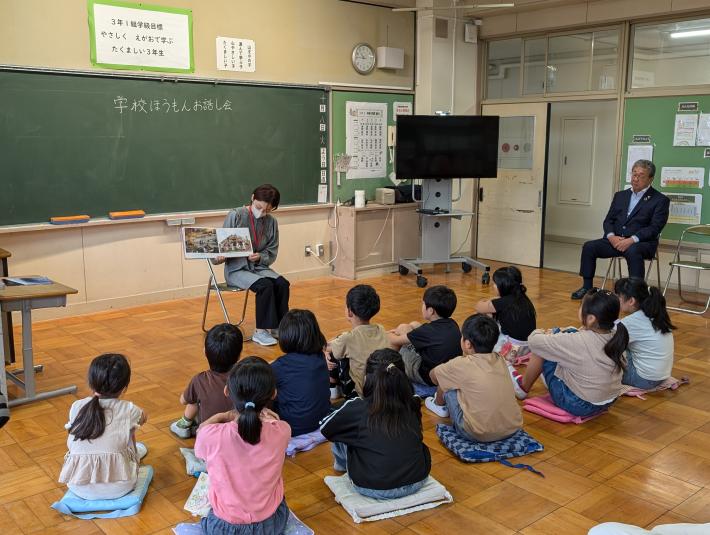 床に座布団を敷いて座る子どもたちが絵本の読み聞かせを聞いている