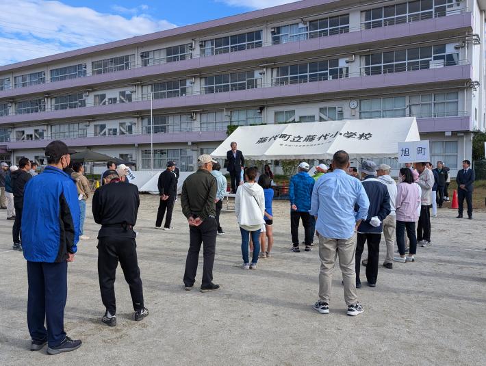 取手市立藤代小学校と書かれたタープの前から、直立の大人たちに向けてあいさつをする男性