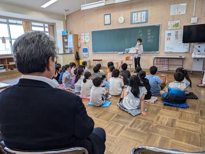 床に座る子どもたちに絵本を読み聞かせる女性。その手前では、男性がいすに座ってその様子を見ている。