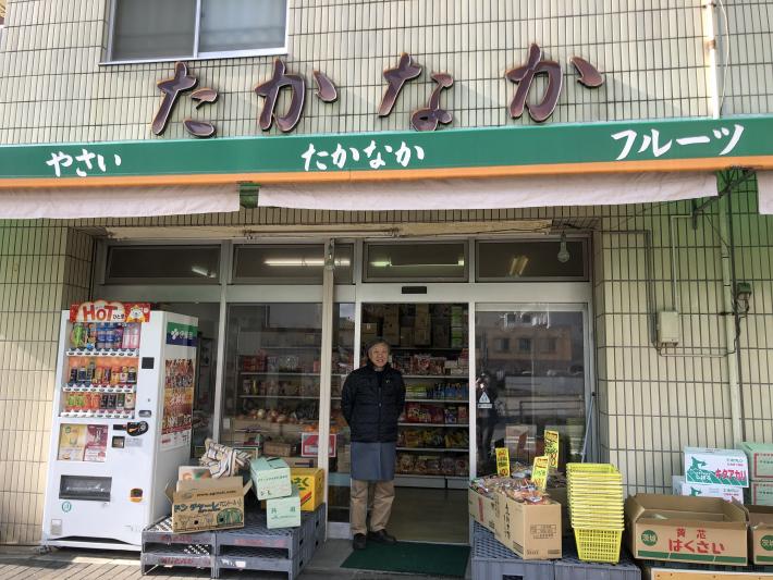 たかなかと書かれた建物に、緑色ののれんがかかっている。店先には、野菜などが並んでいる。