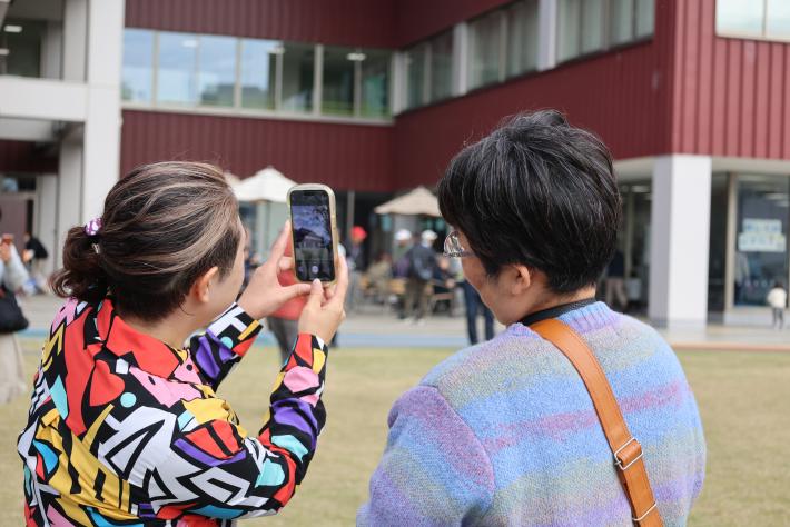 芝生が生えた広場でカラフルな服を着た講師がスマホを構え、そのスマホを青い服を着た女性が見ている