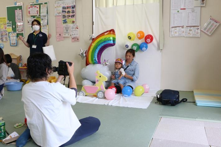 壁にかかった白い布に虹とカラフルな風船が置いてある。布の上に女性と子が座り離れたところで、白いシャツの女性がカメラを構えている