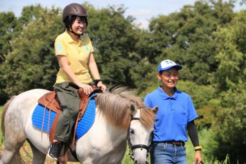 河川敷を乗馬しながらあるく女性とその馬を引く女性